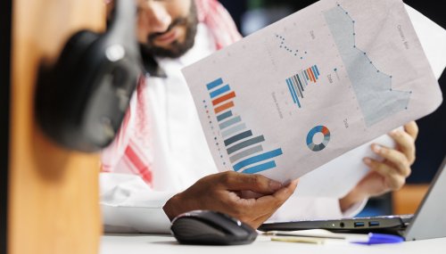 Arab businessman grasping paperwork Focused Muslim man compares information online, surrounded by paperwork on his desk. Selective focus on Arab businessman grasping research papers and conversing on his cell phone with a coworker.